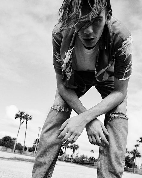 The Road Trip Part 2 - Black and White image of a guy bending over and looking straight down at the camera. He is wearing a short sleeve Hawaiian shirt, open over a white t-shirt and denim jeans with splits on the knees. The Kooks, Jackets Summer, White Image, 2024 Collection, Creepers, Bending, All Saints, Summer Essentials, Leather Jackets