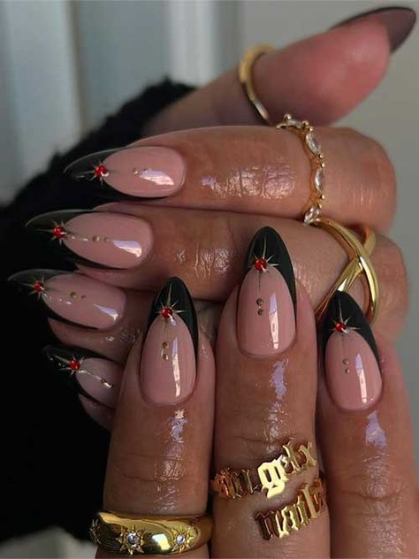 Black French tip nails on a nude base, adorned with gold stars and red rhinestones, featuring gold dots on the nude area. French Tip Nails With Design Rhinestones, Nail Designs Trending Now French Tip, January Nails With Designs, French Tip Color Acrylic Nails, Black And Gold Nails Stars, French Tip Acrylic Nails Glam, Gold Red Black Nails, Black Nails New Year, Almond Black Tip Nails