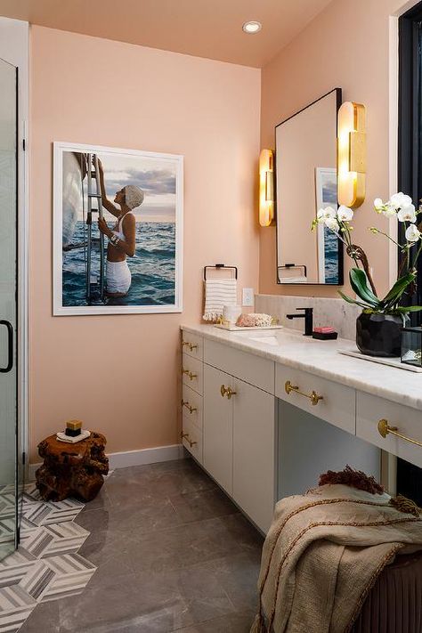 Bathroom features a white bath vanity with brass knobs and a gray honed marble countertop on a pink wall under a mirror lit by brass oval bath lights and gray floor tiles. Gray Floor Tiles, Black French Doors, Terracotta Design, White Bath Vanity, Oval Bath, Gold Pulls, Peach Bathroom, Bamboo Roman Shades, Glam Bathroom