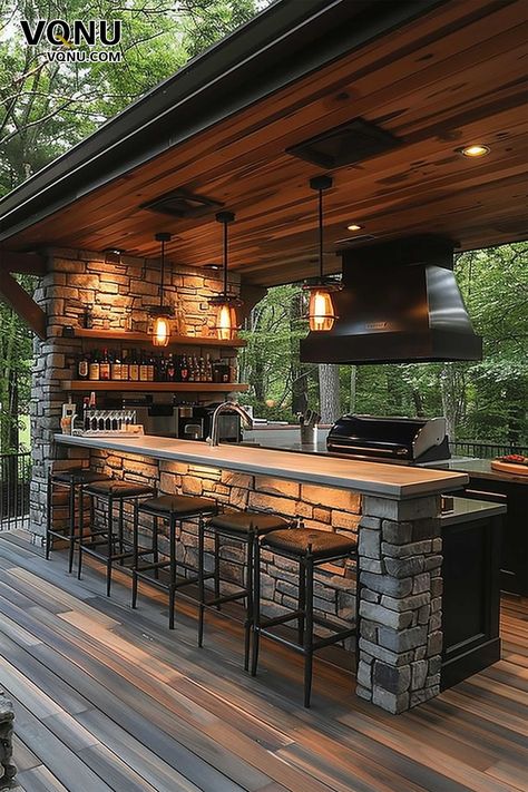 A rustic outdoor bar and kitchen featuring a stone counter, wooden ceiling, and warm lighting. The setup creates a cozy atmosphere, perfect for evening gatherings in a natural, wooded setting. Modern Rustic Outdoor Kitchen, Open Bar Design Outdoor, Modern Outdoor Bar Ideas Backyards, Outdoor Food Setup, Deck With Bar Counter, Covered Patio Bar, Enclosed Outdoor Kitchen, Outside Bar Ideas, Stone Bar Counter