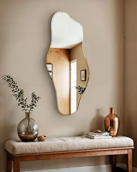 The image showcases the Abstract Wave Wall Mirror, a modern mirror with an intriguing irregular shape that adds a touch of artistic flair to the room. The mirror is mounted above a wooden bench with a soft, cushioned top, adorned with decorative items including a large glass vase with greenery, a metallic vase, and a small stack of books. The overall decor creates a warm and inviting ambiance, blending contemporary design with a cozy, natural feel. Irregular Shape Mirror, Mirror For Living Room, Shape Mirror, Unique Mirror, Wave Wall, Hallway Inspiration, Unique Mirrors, Abstract Waves, Living Room Mirrors