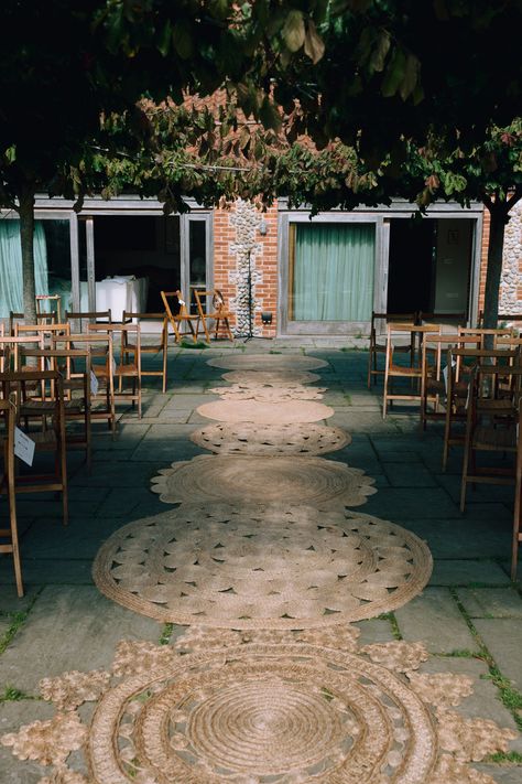 This shows an image showing a ceremony area at an outdoor wedding. On the aisle are different jute rugs lined to accentuate the aisle. Alongside the aisle are vintage folding chairs. Isle Rugs Wedding, Rugs Isle Wedding, Jute Rug Aisle Runner Wedding, Rugs For Outdoor Wedding, Boho Rug Aisle Wedding, Round Rug Wedding Ceremony, Rugs At Wedding Ceremony, Rugs Aisle Wedding, Outdoor Wedding Rugs