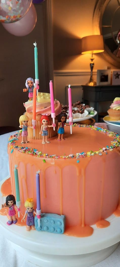 Three tier iced buttercream cake, pink frosting and orange white chocolate gnache drip with lego girls holding multicolored birthday candles! Lego Princess Cake, Lego Themed Birthday Party Girl, Lego Friends Birthday Party Ideas, Lego Birthday Party Girl, Lego Birthday Cake Girl, Lego Friends Birthday Cake, Birthday Cake 7th Girl, Girl Lego Birthday Party, Lego Friends Party Ideas