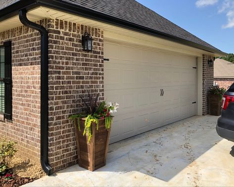 Added black gutters and magnetic hardware to the gatage door. Built the tall planters and stainedto match the front doors and columns ❤️ Brown Brick Black Shutters, Black Gutters Tan House, Black Shutters Brick House, Black Gutters Red Brick House, Black Gutters Gray House, Tan House Exterior, Shutters Brick House, Black Gutters, Garage Colors