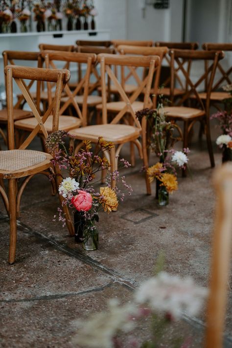 Small floral arrangements in bright colours with retro vibe sit beside wooden chairs lining the aisle | Wedding Aisle Centerpieces, Affordable Wedding Aisle Decorations, Wild Flower Wedding Chairs, Top Of Aisle Decor, Inexpensive Aisle Decorations Wedding, Simple Isle Decoration, Minimal Isle Decor, End Of Aisle Arrangements, Chair Decor Ideas Wedding