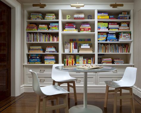 Fantastic game room features wall of built-in bookshelves filled with board games and books illuminated by antique brass picture lights situated behind glass-top game table surrounded by white plastic chairs Library Game Room, White Plastic Chairs, Board Game Room, Garage Game Rooms, Homework Room, Basement Games, Game Room Kids, Board Game Storage, Game Room Basement