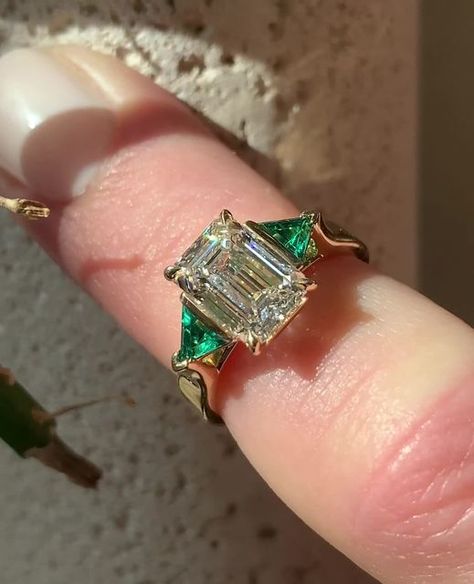 Frank Darling Custom Rings on Instagram: "Send this to someone who loves gemstone accents 💎💚⁠ ⁠ This emerald three stone engagement ring is all custom from head to toe. Let's break it down:⁠ ⁠ • Lab grown green emerald trillion side stones⁠ • 2.1ct, D Color, VVS2 Center Emerald⁠ Diamond • 3.5mm Square band that tapers into cathedral shoulders⁠ • Our signature Breezy setting, to top it off ✨⁠ • All set in a shiny 18k Yellow Gold⁠ ⁠ Mark your calendars, darlings! The last day for Christmas proposal orders is December 1st 💍❄️⁠ ⁠ #emeralddiamondengagementring #emeraldengagementring #threestonering #threestoneengagementring #emeraldcutdiamond #engagementringinspiration #engagmentringideas #gemstonering #customengagementring⁠ ⁠ 𝐖𝐚𝐧𝐭 𝐭𝐨 𝐠𝐞𝐭 𝐬𝐭𝐚𝐫𝐭𝐞𝐝 𝐝𝐞𝐬𝐢𝐠𝐧𝐢𝐧𝐠 𝐲𝐨𝐮𝐫 ? Diamond Ring Emerald Band, Diamond Ring With Colored Side Stones, Engagement Rings Diamond And Emerald, Engagement Ring With Emerald Side Stones, Engagement Ring Diamond And Emerald, Emerald Accent Engagement Ring, Yolanda Diaz Wedding Ring, Emerald Side Stone Engagement Ring, Vintage Setting Engagement Ring