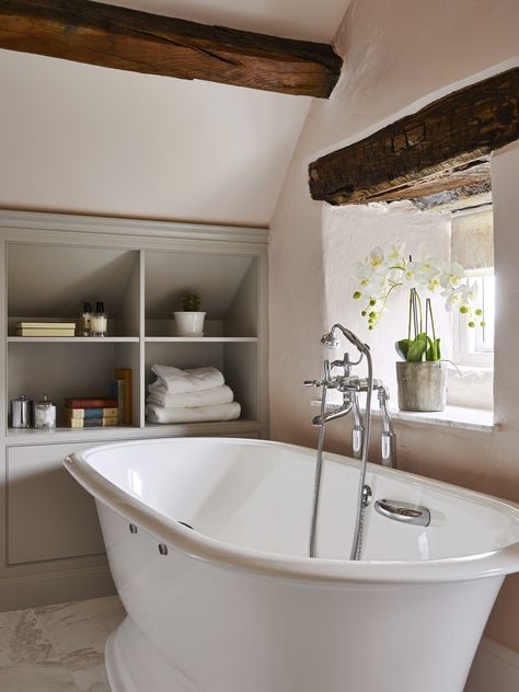 Sloping ceilings and heavy beams can present design challenges in older properties. The bathroom featured was designed to maximise storage with cabinetry and shelving handmade to fit the available space under the eaves. Freestanding bath from @vandabaths, taps and accessories from @samuel_heath and furniture finished in #purbeckstone from @farrowball.⁠ ⁠ #bathrooms #ensuite ⁠ #bathroom #bathroomdesign ⁠ #bathroomcabinetry #bath ⁠ #bathroomdesigners ⁠ #bespokebathroom⁠ #yorkshirebathroom⁠ Eaves Bathroom Storage, Bathroom Eaves Storage, Bathroom Sloping Ceiling, Bathroom In The Eaves, Shower Under Sloped Ceiling, Bathrooms With Sloping Ceilings, Exposed Beams Bathroom, Bathroom With Eaves, Luxury Cottage Bathroom