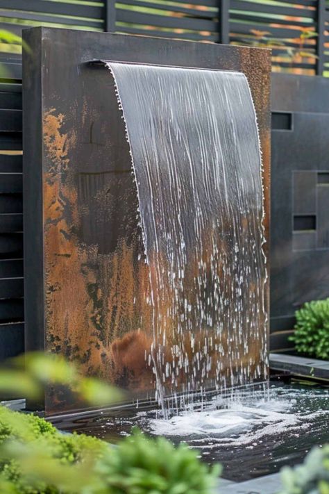 A rustic metal wall fountain with water gracefully flowing over a textured surface, surrounded by greenery in a stylish garden setting. Garden Wall Fountains Outdoor, Diy Outdoor Waterfall Fountain, Outdoor Wall Fountains Modern, Water Features In The Garden Fountain, Mini Fountain Garden, Water Falls Backyard, Wall Waterfall Outdoor, Garden Feature Wall, Backyard Fountain Ideas