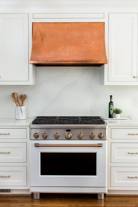 Fixed between white inset cabinets, a copper range hood is mounted against a honed marble backsplash and over a white stove with brass trim flanked by white cabinets topped with a honed marble top. White Kitchen Vent Hood, White Stove, Montgomery Homes, Kitchen Island Tops, Inset Cabinetry, Kitchen Vent Hood, Copper Hood, Light Gray Cabinets, Kitchen Vent