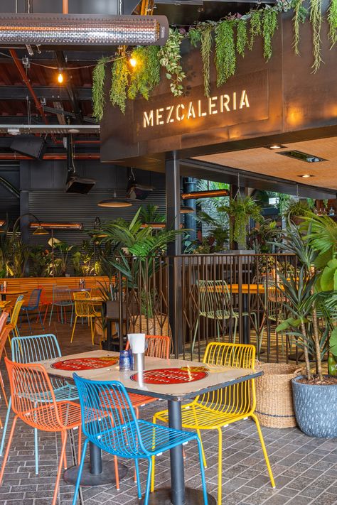 bright coloured seating area with square glossy wooden table, floral background with a mixture of different plants creating a beautiful atmosphere. big open plan restaurant and bar area with black walls and stone floor. Mexican Restaurant Outdoor Patio, High End Mexican Restaurant, Aesthetic Mexican Restaurant, Mexican Cafe Design, Mexican Restaurant Patio, Modern Mexican Restaurant Design, Mexican Bar Ideas, Mexican Restaurant Interior Design, Outdoor Restaurant Ideas