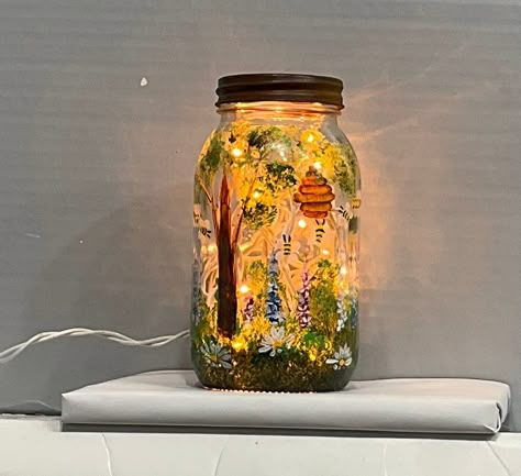 A clear glass quart canning jar was recycled to make this charming light. It can be used as a night light in any room. It will add unique mood lighting to any room. The quart jar was drilled in the back side and a strand of 20 lights was inserted through this hole so that the cord comes out the side leaving 24 inches of cord length to reach an outlet. It was then hand painted by myself using enamel glass paint. Size 7 inches high The painting is of a garden, flowers, tree, bee hive and bee Hand Painted Mason Jars, Mason Jar Decorating, Glass Jar Lamp, Mason Jar Painting, Mason Jar Light, Painting Glass Jars, Lighted Glass Blocks, Painted Glass Bottles, Glitter Mason Jars