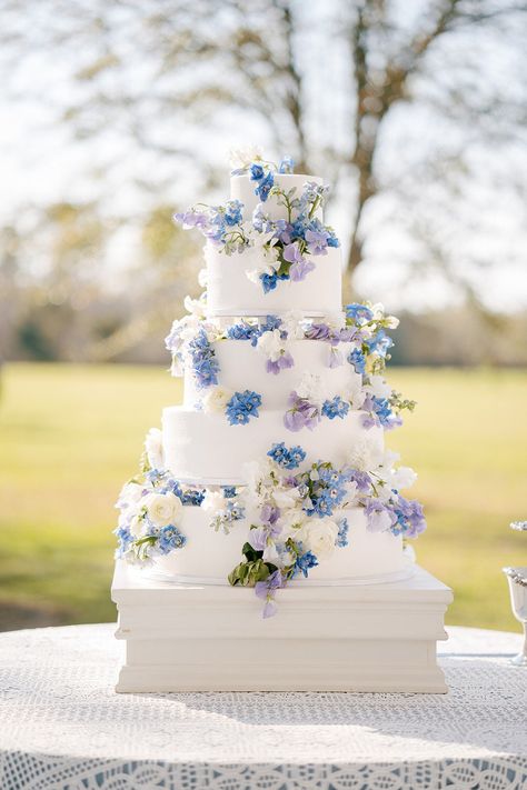 Multi-tier wedding cake decorated with blue, white and purple flowers. Photo: @alishacrossleyphotography Dusty Blue And Lilac Wedding Decorations, Pastel Blue And Lavender Wedding, Lavender And Blue Wedding Cake, Wedding Themes Blue And Purple, Purple And Blue Wedding Cake Ideas, White Blue And Purple Wedding, Pastel Blue And Purple Wedding Theme, Light Blue Lavender Wedding, Wedding Cakes Blue And Purple