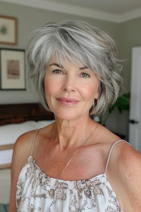 Smiling woman with short grey hair in a casual bedroom setting. New Gray Hair Trend, Haircuts 2024 Medium Hair, Shaggy Gray Hairstyles, Gray Hair Wigs, Short Grey Hair With Lowlights, Medium Silver Hairstyles, Chin Length Gray Hairstyles, Gray Hair Shoulder Length, Over 60 Grey Hairstyles For Women