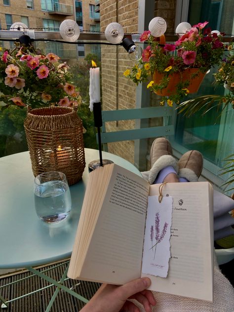 Girl sitting with her feet up reading her book with a handmade watercolor bookmark in on her balcony. The balcony is full of plants and flowers and the sun is setting in the background. Candles are lit and it’s the perfect ending to a day. Reading On Balcony Aesthetic, Slow Living In The City, Reading On Balcony, Books And Plants Aesthetic, Balcony Candles, Slow Aesthetic, Reading Balcony, Reading In The Garden, Live Slowly