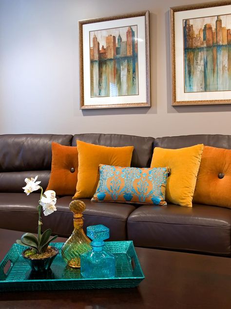 A brown leather sofa matches a dark wooden coffee table in front of the neutral living room walls. Orange throw pillows and a contrasting patterned blue pillow add color to the room. Two scenic photos and a bright blue breakfast tray pull repeat the hue. Leather Couch Decorating, Living Room Decor Orange, Sofa Kulit, Blue Couch Living Room, Brown Sofa Living Room, Turquoise Room, Brown Theme, Brown Living Room Decor, Brown Couch Living Room