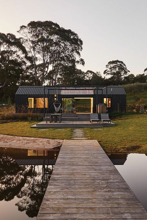 The Agricultural Lakehouse is an off-grid retreat. Sitting amongst rugged bushland with a blazing sunset in the background, the modular home is sitting on the banks of a picturesque dam that reflects the amazing surroundings. Clad in a deep charcoal Colorbond with black trim, pergolas and window frames. Prefab Container Homes, Off Grid House, Shed Home, Prefab Cabins, Landscape Construction, Prefabricated Houses, Shed Homes, Container Homes, Shipping Container Homes