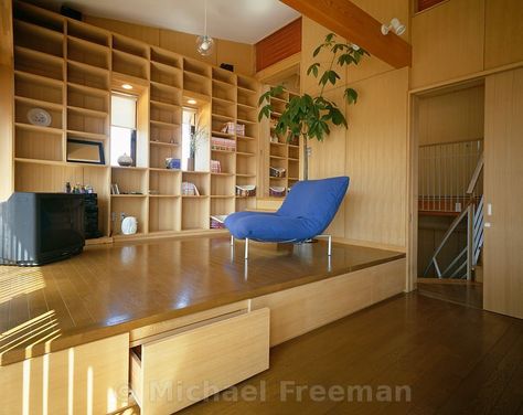 The living room of a low-cost Japanese home in Chiba, Japan, by architect Shintaro Hanazawa. The sitting area is on a raised wooden platform that provides additional storage space, with a grid of wooden shelving covering one wall. Raised Wooden Platform Living Room, Raised Platform Living Room Floors, Platform Living Room Raised, Raised Floor Storage, Japanese Reading Room, Raised Floor Living Room, Raised Living Room Floor, Japan Tiny Apartment, Raised Platform Living Room