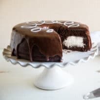 A square photo of the homemade ding dong cake taken from the side of the cake. There is a slice taken out of the cake and the creme filling is exposed. The white squiggle frosting is visible on the top of the cake, on top of chocolate frosting that drips down the side of the cake. The cake is on a white cake stand that has wave-like edges. It is taken against a white background. Pie, Homemade Ding Dongs, Chocolate Marshmallow Cake, Ding Dong Cake, Creme Filling, Marshmallow Filling, Hot Milk Cake, Kid Friendly Dessert, Rich Chocolate Cake