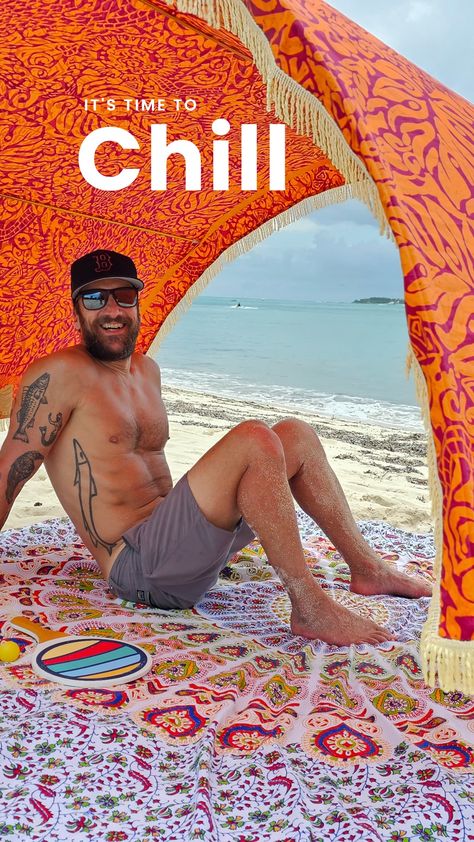 A man is sitting beneath an orange, arched shade cabana.  He is wearing grey shorts, his muscular upper body is shirtless, and he is smiling.  The turquoise ocean is behind him, and he sits on a colourful cotton beach mat. Beach Cabanas, Beach Tents, Portable Shade, Beach Cabana, Windy Weather, Beach Tent, Carry On Suitcase, Beach Travel, The Cool