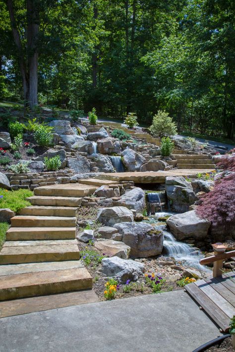 Modern Design Aquascaping did a tear-out & re-installation of a pondless waterfall in Lenoir City, TN. The feature which was done by Modern Design Aquascaping, includes large mountain boulders,cedar driftwood accents, LED under-water lighting, flagstone walkway-steps-and bridge, and a wooden bridge. Cascade Waterfall Garden, Backyard Creek Landscaping, Mountain Garden Ideas, Pondless Waterfall, Flagstone Walkway, Fountains Backyard, Sloped Backyard, Hillside Landscaping, Backyard Water Feature