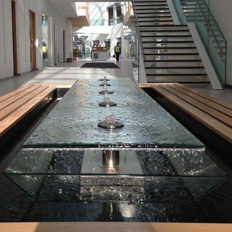 A stunning water feature. The design has multiple concentric rings of water that are brought to life with the light through the glass.  Water falling like rain around the outside gives another dimension as well as sound. Residential Water Feature, Luxury Water Feature, Indoor Water Features Living Rooms, Lobby Water Feature, Lobby Reception Design, Indoor Waterfall Wall, Garden Water Features, Reception Area Design, Indoor Water Features