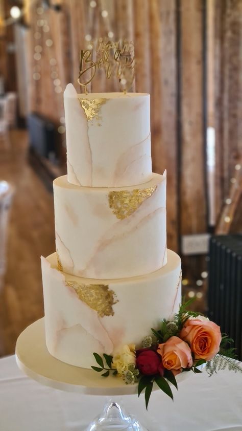 Three tier torn paper effect wedding cake with nude and blush dusting and edible gold leaf accents, sitting on clear glass cake stand with fresh flowers on the bottom tier in a barn setting with beams and fairy lights in the background Wedding Cake Champagne Color, Blush And Gold Wedding Cake, Honey Bee Wedding, Wedding Cake Gold Leaf, Bee Wedding Cake, Wedding Cake Two Tier, Torn Paper Effect, Wedding Cake Marble, Cakes With Flowers