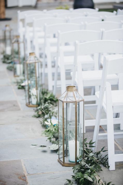 Aisle in ceremony lined with greenery and lanterns with candles Gold Lantern Aisle Decor, Lanterns Lining Wedding Aisle, Greenery Lined Wedding Aisle, Sage Green Isle Decor, Sage Wedding Aisle Decor, Wedding Aisle Decorations Outdoor Lanterns, Beach Wedding Lanterns Aisle, Sage Green Isle Wedding, Wedding Aisle Lanterns Outdoors