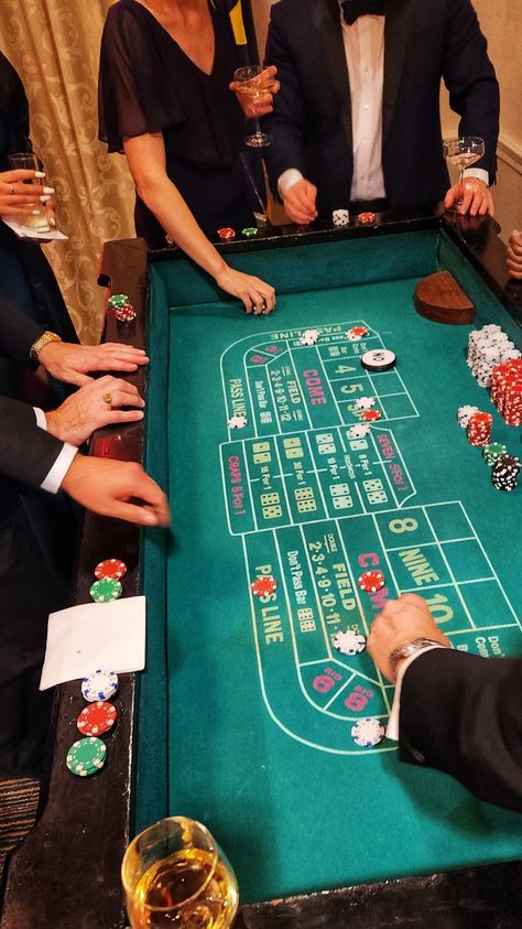 A craps table surrounded by tux covered hands, wine glasses, ladies in evening gowns. Wine glass in the lower corner, dice and chips on the table as play is in session Las Vegas, Craps Table, Imaginative Writing, Casino Room, Surprise 60th, Green Room Decor, Casino Birthday, Casino Table, Poker Party