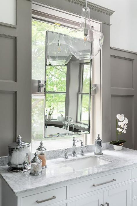 A tall vanity mirror in front of a bathroom window sits over a white washstand with gray marble countertop and a nickel hook and spout faucet illuminated by a Kichler pendant. Paneled Mirror Bathroom, Mirrors In Front Of Windows Bathroom, Bathroom Mirror In Window, Bathroom Mirror In Front Of Window, Mirror In Front Of Window Bathroom, Mirror In Front Of Window, Vanity In Front Of Window, Berry Bathroom, Laundry Combo