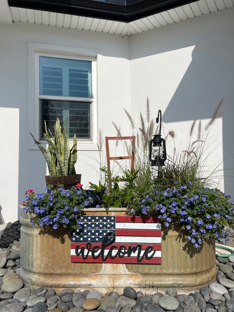 A variety of zone, sunlight, and soil plants inside a livestock tank with added visual interest such as a rustic bucket, a ceramic hen nestled in the plants, a small shepherd’s hook with an antique lantern, and a patriotic sign hanging on the front. At night, the display, which sits between the driveway and front door, is lit up by a solar spotlight. Repurposed Water Trough, Water Trough Ideas Backyards, Stock Tank Repurposed, Cow Trough Garden, Trough Flower Planters, Trough Flower Garden, Water Trough Flower Planter, Metal Horse Trough Planters, Old Water Trough Ideas