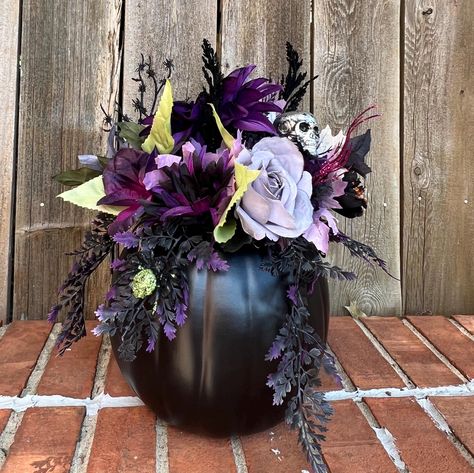 This  flower arrangement features all the things that make Halloween fun and  beautiful.  The black artificial pumpkin has the lid carved out and attached to the back of the arrangement.  The center is filled with flowers in the hues of gray, purple, black, and green.  Fun picks feature a silver skull, a purple and a green spider small spider web.  Black and purple feathery sprays and glittery berry sprays complete this arrangment   The pumpkin arrangement measures 15" in heigth and 12" at the widest part.   Item #FLR - 84 Spooky Flower Arrangements, Spooky Apartment, Flower Diy Paper, Halloween Flower Arrangements, Quince Planning, Halloween Floral Arrangements, Paper Flower Diy, Green Spider, Fall Pumpkin Centerpieces