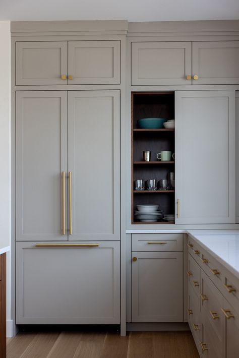 A streamlined kitchen corner boasts taupe Shaker style cabinetry with gold hardware, and chic white marble countertops. Cabinet doors slide to expose custom built-in shelves for easy organization, and the hidden refrigerator adds a sophisticated touch. Hidden Appliances, Coastal Color Scheme, Gold Kitchen Hardware, Modern Shaker Kitchen, Yarmouth Maine, Gray Doors, Taupe Kitchen, Shaker Style Kitchen, Shelving Kitchen