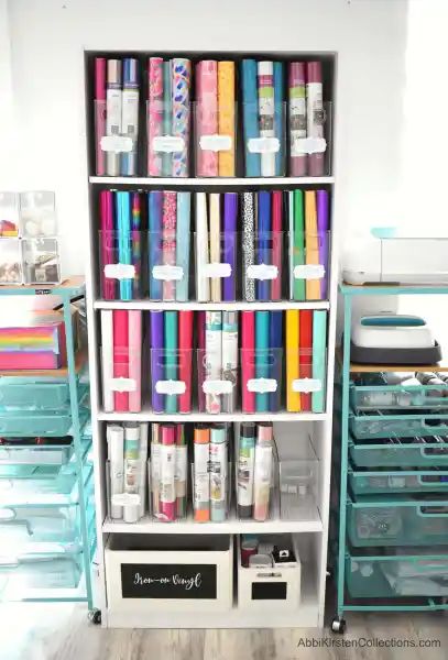 A white, tall bookshelf holds Cricut vinyl in clear storage bins organized by type. Labels make finding what you need easy and hassle-free. Organizing Vinyl Rolls, Vinyl Roll Storage Ideas Diy, Craft Room Printables, Storage For Craft Room, Cricut Office, Craft Storage Ideas For Small Spaces, Office Craft Room Combo, Cricut Storage, Organizing Office