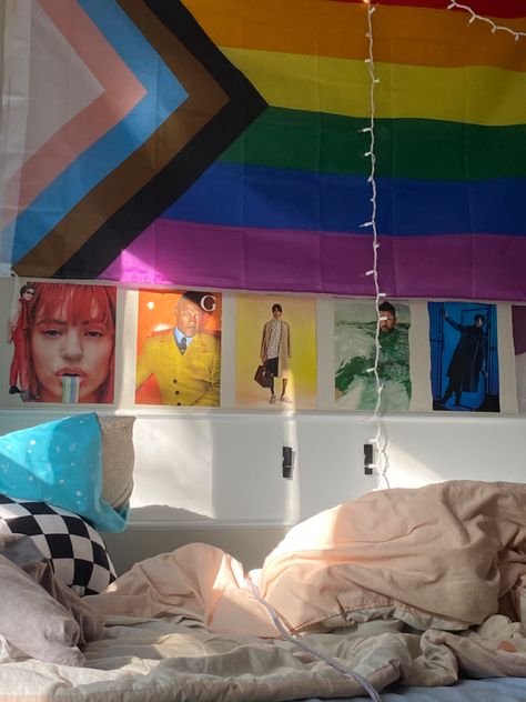 a picture of a room, featuring a bed with a light pink blanket, a small black and white checkered pillow, and a light blue pillow. a wall is decorated with a pride flag, and rainbow magazine cut outs beneath it. fairy lights hang from the wall as well. Lgbtq Room Ideas, Pride Flag In Room Aesthetic, Lesbian Flag Room, Pride Flag Bedroom, Pride Flag Room Decor, Pride Flag In Bedroom, Lgbtq Room Decor, Flag Display, Rainbow Fairies