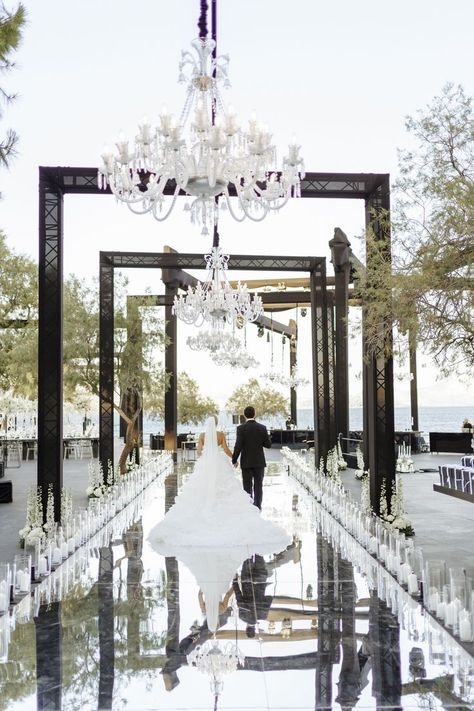 Newlyweds while walking down the aisle of their wedding venue. An aisle made of mirrors and decorated with plenty of candles, some white flowers and chandeliers. Classy Outside Wedding, Island Art And Taste Wedding, Modern Wedding Venue Ideas, Monochromatic Wedding, Wedding Aisle Outdoor, Athens Riviera, White Wedding Ceremony, Monochrome Weddings, Black And White Wedding Theme