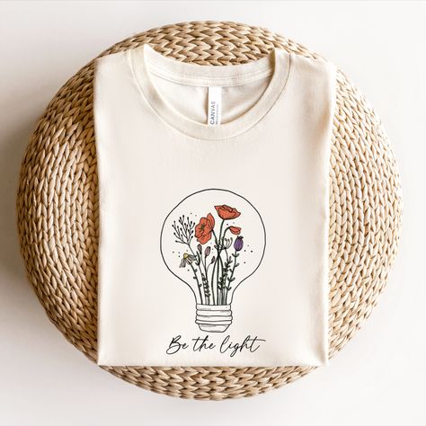 Floral shirts for women, Spring Shirt. This design with the quote "Be the light" and a lovely wildflower growing inside a light bulb is the perfect way to inspire yourself and others to shine bright and make a positive impact on the world. The beautiful illustration features a wildflower growing inside a light bulb, reminding you to nurture your own inner light and let it shine. The quote "Be the light" is a powerful reminder to be a positive influence and make a difference in the world. Roots Clothing, Light Flowers, Quote Graphic, Floral Shirts, Positive Influence, Be The Light, Inspiration Quote, Beautiful Illustration, Lighted Canvas