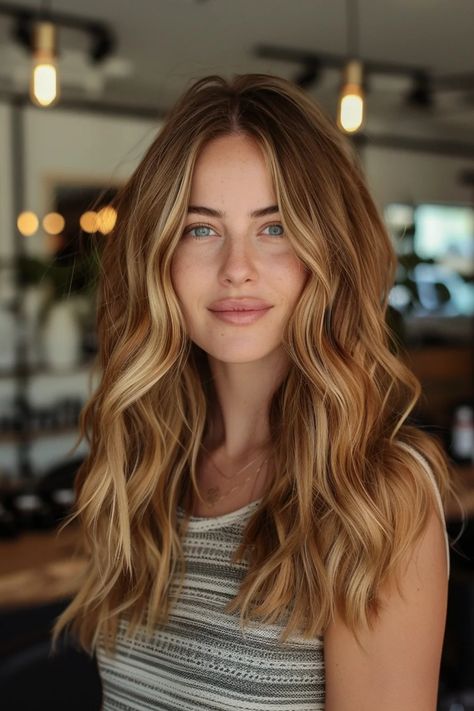 Woman with long wavy blonde hair and a striped top smiling indoors with blurred lights in the background. Caramel Hair Balayage Honey, Brown Hair Base With Blonde Highlights, Balayage, Caramel On Blonde Hair, Light Brown Hair With Caramel And Blonde Highlights, Dark Blonde With Highlights Sun Kissed, Light Brown Hair With Golden Undertones, Light Caramel Brown Hair With Highlights, Golden Fall Hair