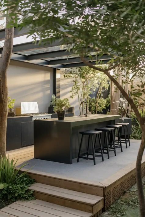 Outdoor kitchen with a sleek black bar, bar stools, and lush greenery, under a modern pergola. Concrete Outdoor Kitchen, Fire Pit Seating Area, Bar Counter Design, Industrial Modern Design, Black Stool, Island Stools, Island Chairs, Modern Outdoor Kitchen, Black Kitchen Island