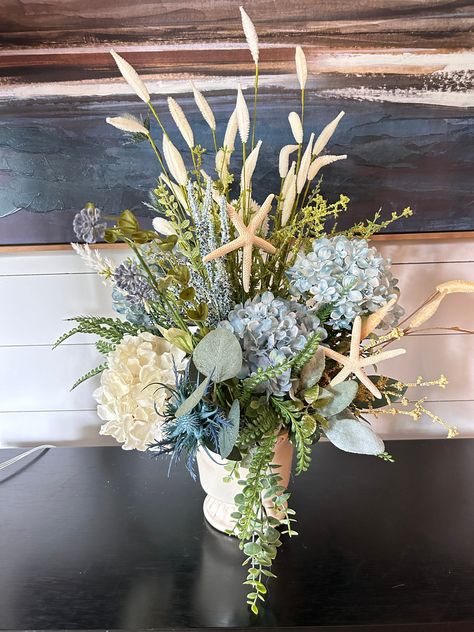 This coastal flower centerpiece is beautiful!   It is made with all artificial pale blue and cream hydrangeas and stems of coastal style foliage and greenery. Natural preserved starfish add a classic coastal touch.  This pretty nautical centerpiece is arranged in a beige ceramic vase, florals are permanently attached inside of the vase.  All sides are displayable.  Measures at about 22 inches high at the tip of the florals, 12-14 inches in diameter at widest points at the greenery edges.  Vase i Ocean Centerpieces Wedding, Beach Theme Flower Arrangements, Beachy Centerpieces, Coastal Centerpiece, Nautical Centerpiece, Beach Centerpieces, Beach Wedding Centerpieces, Beachy Wedding, Hydrangea Centerpiece