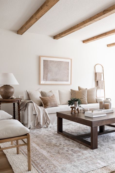 The space is elegantly adorned with a subtle blend of natural textures and soft neutrals, highlighted by the rustic charm of wooden ceiling beams. A plush off-white jute rug anchors the room, complementing the earthy tones of the accent pillows and the rich, dark wood coffee table. The delicate balance of comfort and style is perfected by a soft throw, an artful ladder, and a statement ceramic lamp, all inviting a sense of calm and simplicity. White Sofa Area Rug, Living Room Rugs White Couch, White Jute Rug Living Room, Dark Wood And White Living Room, White Couch Rug Ideas, Neutral Living Room Furniture, Affordable Living Room Decor, Dark Wood Coffee Table, Jute Rug Living Room