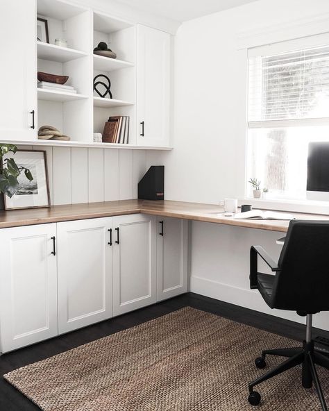 A spacious office with an L-shaped desk built into one wall features a black-and-white color scheme and a minimalist design. A woven rug covers the floor and a simple black office chair sits on top in front of a computer monitor and an open window with white blinds. Cubed shelves hold books, framed images, wooden baskets, and other modern decor. A plant, a larger print, and a small black magazine organizer adorn the desk. L Shaped Office Desk Ideas Layout, Built In L Shaped Desk, L Shaped Office, Built In Desk And Shelves, Home Office Built Ins, L Shaped Office Desk, Spacious Office, Office Built Ins, Minimalist Home Office