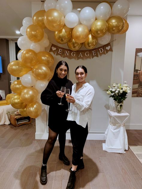 Two Indian women standing in front of decoration display made up or gold and white balloons and a banner that says engaged on it, cheersing with champagne glasses Black White Gold Engagement Party, Simple Engagement Party Ideas At Home, Casual Engagement Party Decorations, Small Engagement Party Ideas Simple, Gold Engagement Party Decor, Simple Engagement Decorations, White And Gold Engagement Party, Engagement Party Decorations Indoor, At Home Engagement Party