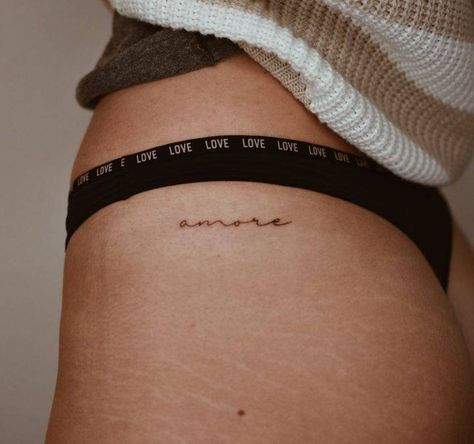 Tattoo of the word "amore" located on the hip. Amor Tattoo On Hip, Small Tattoos For Women Hip Bone, Amor Hip Tattoo, Words Hip Tattoo, Danty Tattoos Small On Hip, One Word Hip Tattoo, Upper Thigh Writing Tattoo, Hip Area Tattoo, Hidden Hip Tattoo