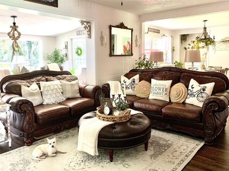 This farmhouse living room boasts a brown leather couch with antique dark wood frame seated next to a matching loveseat. A round tufted brown leather ottoman is placed in front of the couch on a beige and gray vintage rug. Dark Brown Black And White Living Room, Styling A Dark Leather Couch, Farmhouse Living Room Brown Leather Sofa, Brown Loveseat Living Rooms, Dark Brown Leather Couch Living Room Rug, Living Room With Dark Brown Leather Furniture, Decorating With Dark Brown Leather Couch, Dark Leather Furniture Living Room, Pillows For Dark Brown Leather Couch