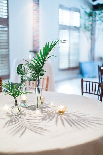 Tropical centerpiece filled with monstera leaf and areca palms. Surrounded with gold votives. Floral by Duvall Events. #duvalleventschs, #tropical, #monstera, #arecapalm, #modern, #charleston, #centerpiece Classy Tropical Centerpiece, Palm Centerpiece Simple, Minimal Tropical Wedding Centerpiece, Tropical Leaves Centerpiece, Palm Leaf Table Decor, Tropical Leaf Centerpiece, Simple Tropical Centerpieces Wedding, Palm Leaf Centerpiece Wedding, Tropical Greenery Centerpieces