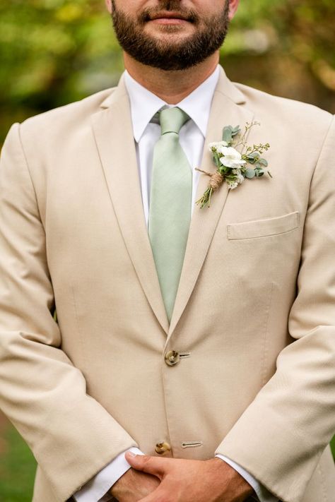 Close up picture of male wearing a tan suit jacket, with white shirt and sage green tie. Small green and white flowers wrapped in twine accent the right lapel Sage Groom, Rustic Wedding Groom Attire, Tan Groomsmen Suits, Tan Suit Wedding, Groom Outfit Inspiration, Easton Maryland, Rustic Wedding Groom, Suit Inspiration, Green Wedding Suit