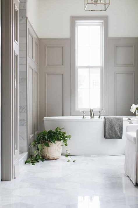 Gray wood paneling accents a wall wall and partially frames a window positioned above an oval freestanding bathtub placed on large marble hex floor tiles. Gray Wainscoting, Paneling Design, Gray Makeup, Bathroom Transitional, Oval Bathtub, Fur Stool, Armoire Design, French Bathroom, Bathroom Paneling