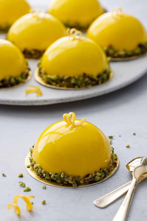 Bright yellow mirror-glazed dome shaped mousse cakes with pistachios around the edges, one cake in front and a plate of more in the background. Mini Dome Dessert, Lemon Shaped Dessert, Mousse Cake Decoration Ideas, Lemon Mousse Cake Recipe, Dome Desserts, Lemon Mousse Cake, Meyer Lemon Curd, Mirror Glaze Recipe, Entremet Recipe