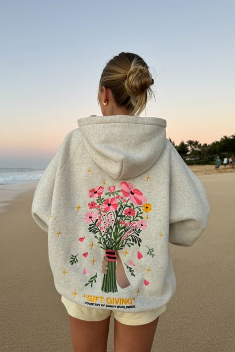 photo shows a blonde teenage girl standing on a sandy beach at sunset. she is facing away from the camera, looking at the ocean in the distance. she is wearing cream colored sweat shorts and a gray hoodie that has a big embroidered graphic of a bouquet of flowers on the back. "gift giving" and "courtesy of dandy worldwide" are embroidered on the back as well. the girl has her hair in a bun. the sky is orange and blue, like the sun is just about to set Dandy Flower Hoodie, You Should Buy Her Flowers Hoodie, Gift Giving Hoodie, Cute Aesthetic Hoodies, Winter Hoodie Outfit Women, Hoodie Embroidery Ideas Boyfriend, Dandy Worldwide Hoodie, Dandy Sweatshirt, Beachy Hoodies